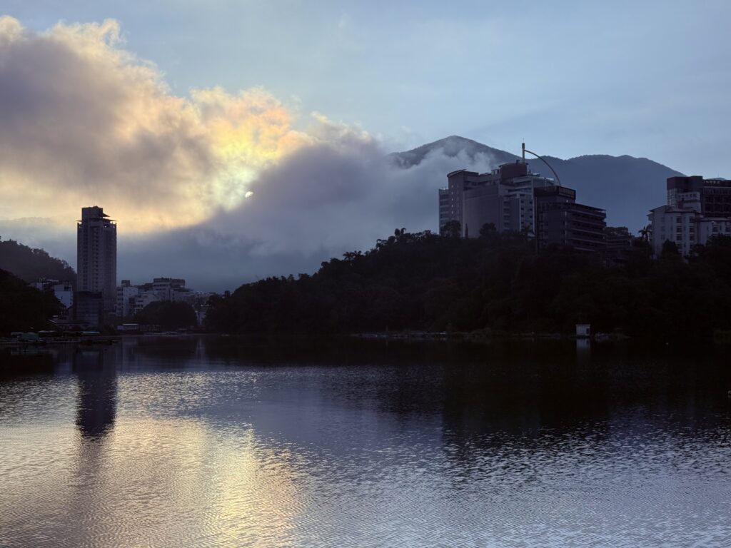 A sliver of the sun, behind clouds, popping out from behind a mountain, shining down on a lake